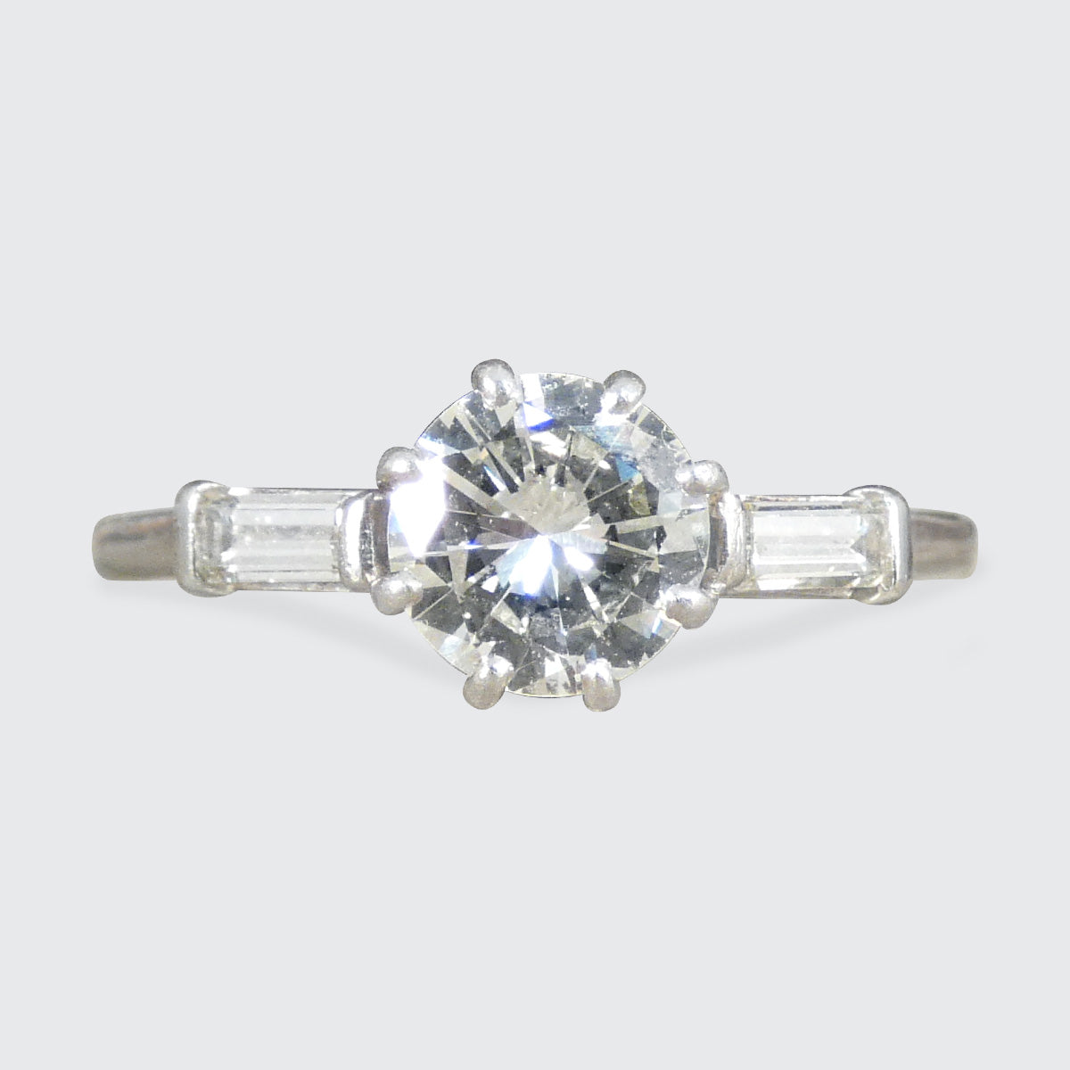 Art Deco platinum ring featuring a dazzling round brilliant-cut diamond as the centrepiece, flanked by elegant baguette-cut diamonds on either side. The design reflects timeless sophistication with a sleek, geometric aesthetic.