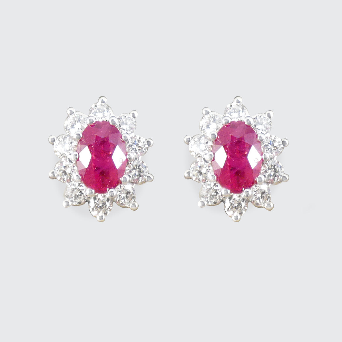 Ruby and Diamond Cluster Stud Earrings in 18ct White Gold, in a classic setting that will stand the test of time. Featuring a bright and pinkish oval cut Ruby in each stud with a cluster of Diamonds in 18ct White Gold.