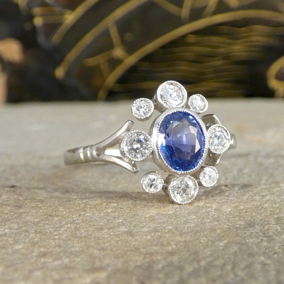 A stunning Sapphire and Diamond ring crafted in platinum, featuring a central vibrant blue sapphire surrounded by a halo of round brilliant-cut diamonds. This ring showcases a unique art deco-inspired design with delicate milgrain detailing, exuding timeless elegance and sophistication.