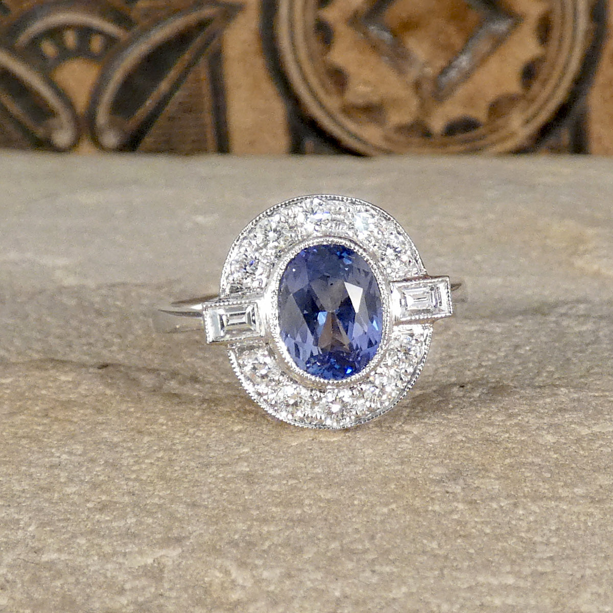 An oval cut Sapphire with a rub over bezel setting surrounded by Brilliant and Baguette cut Diamonds clustering the velvet blue centre stone. A true are deco style ring, a beautiful vintage classic.