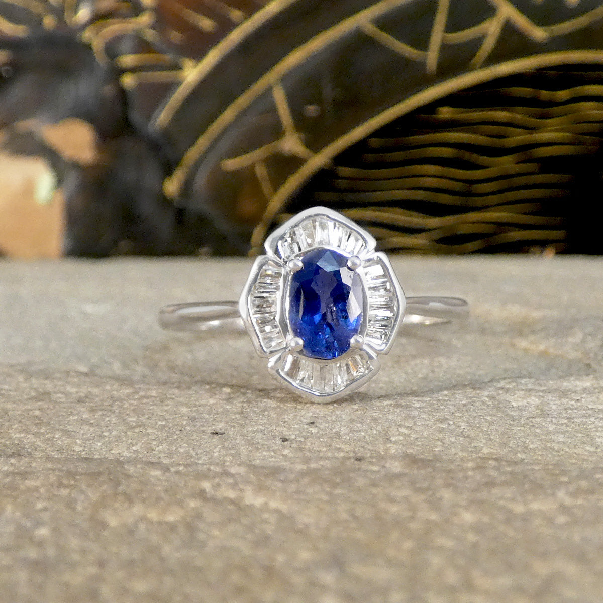 Elegant silver coloured ring with a striking oval blue sapphire centre stone, surrounded by a halo of baguette-cut clear gemstones in a unique, art deco-inspired setting, displayed on a stone surface with a textured, decorative background.