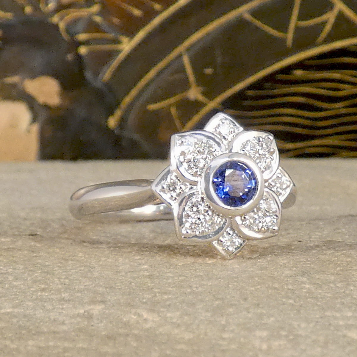 A flower cluster ring featuring a Ceylon Sapphire in the centre in a rub over bezel setting with a petal shaped Diamond clusters all made in Platinum. Showing a bright blue hue.