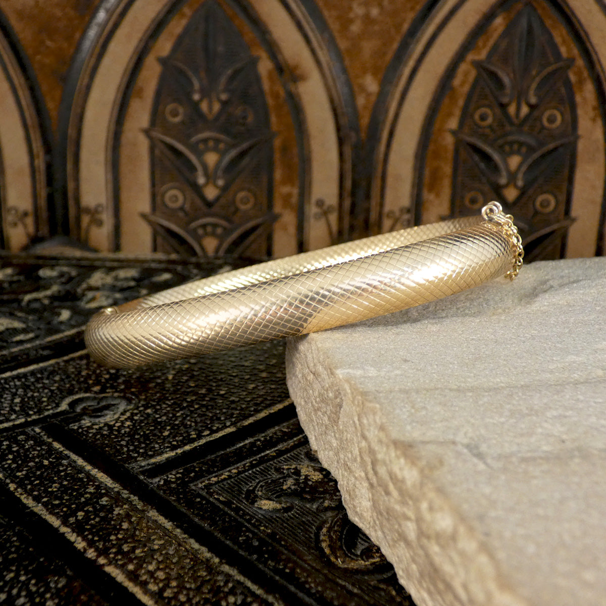 Vintage 9ct yellow gold bangle with a textured design, displayed on a stone surface against a decorative Gothic-style background.