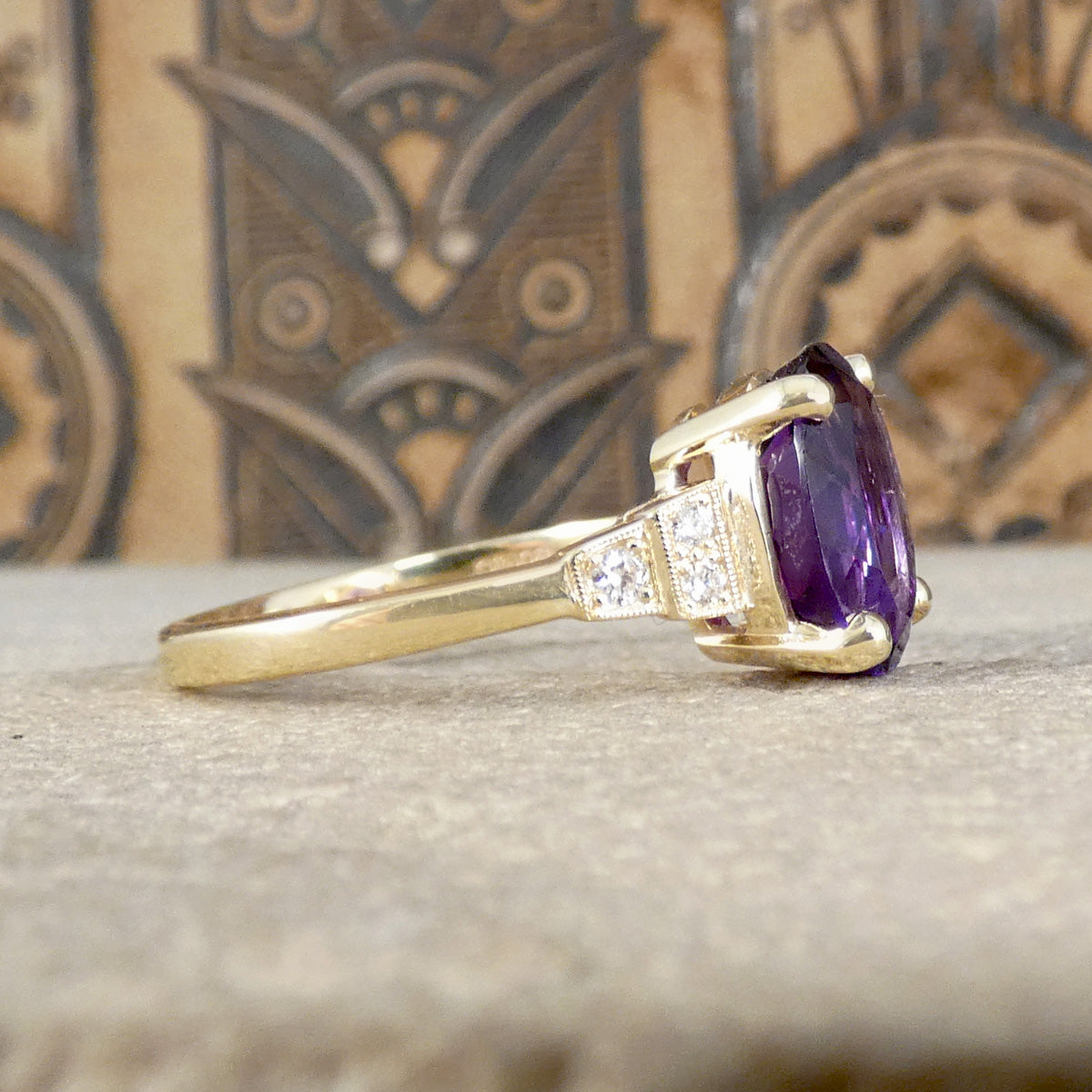 Side view of bright and vibrant amethyst ring in a four claw setting with diamond set shoulders in 9ct yellow gold, showing milgran detail in setting.