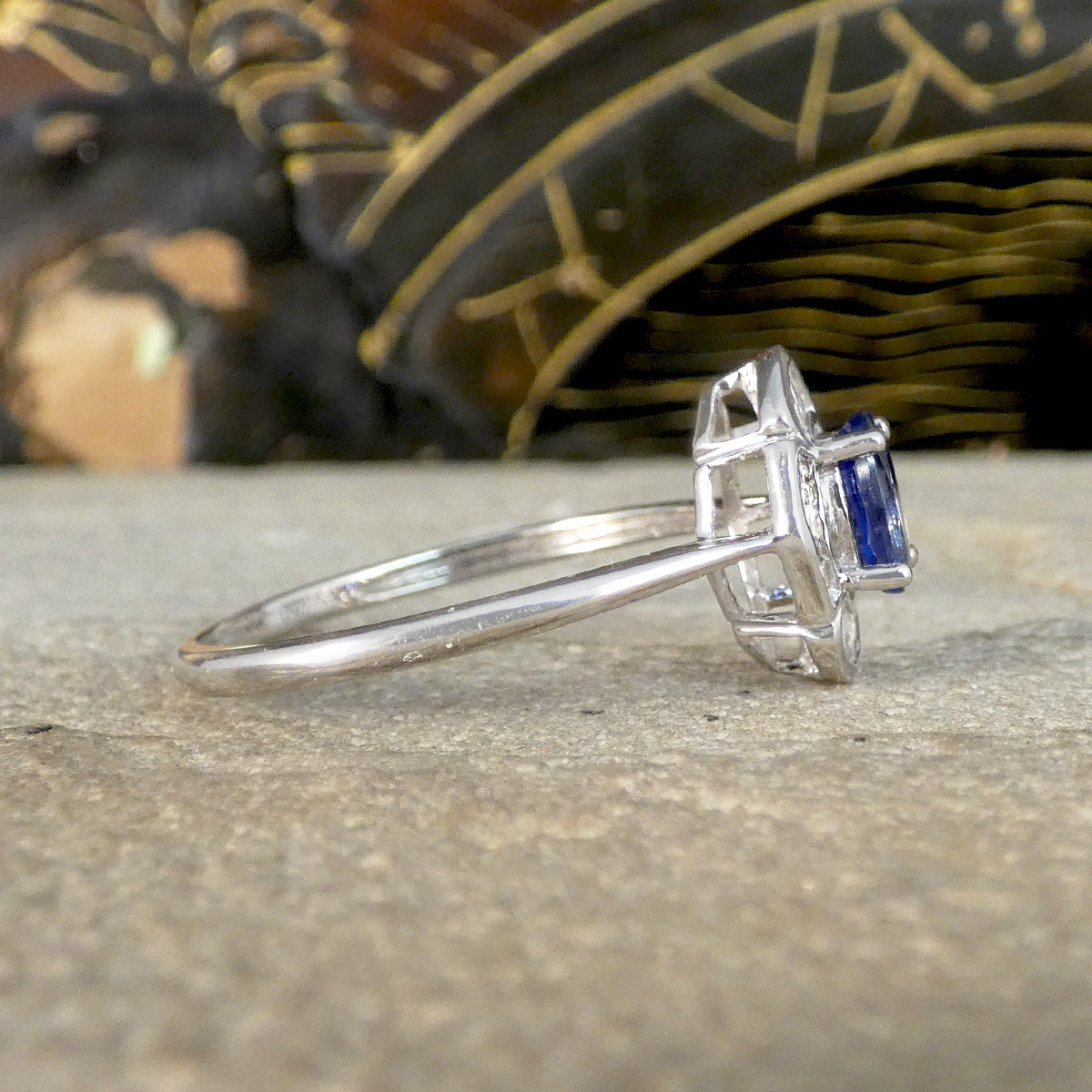 Side profile view of a White Gold ring featuring an oval blue sapphire centre stone, showcasing the elevated, intricate setting with a geometric design, highlighting the craftsmanship and unique structure against a stone background.