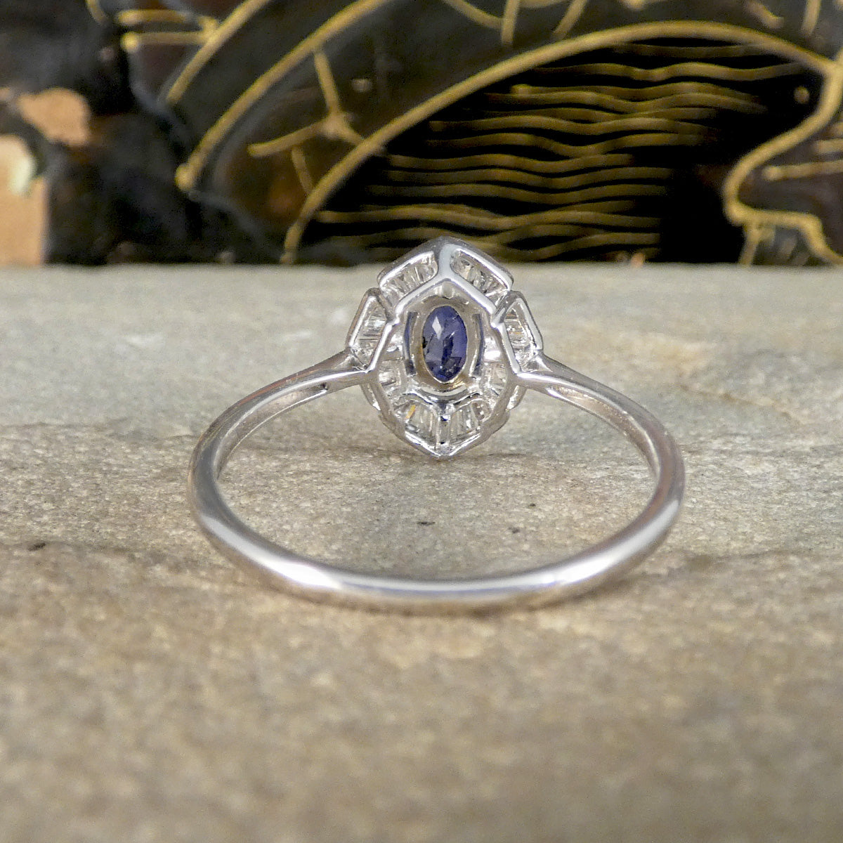 Back view of a White Gold ring with an oval blue sapphire centre stone, showing the intricate, art deco-inspired setting with clear baguette accents and a slim, polished band, against a stone-textured background.