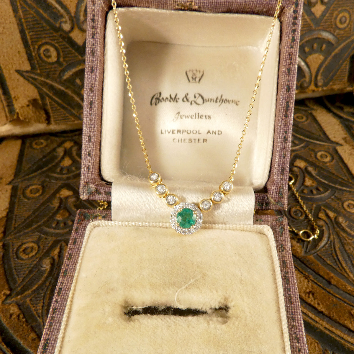 A stunning new necklace crafted in 18ct yellow gold, featuring a vibrant green emerald centre piece surrounded by a halo of sparkling diamonds, complemented by additional bezel-set diamonds in an illusion setting along the chain. The perfect gift. 