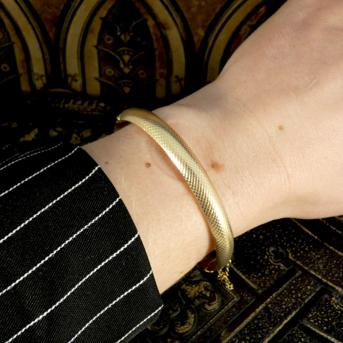 Vintage 9ct yellow gold hollowed bangle with a textured design, shown worn on a wrist, paired with a black pinstripe outfit and ornate Gothic-style background.