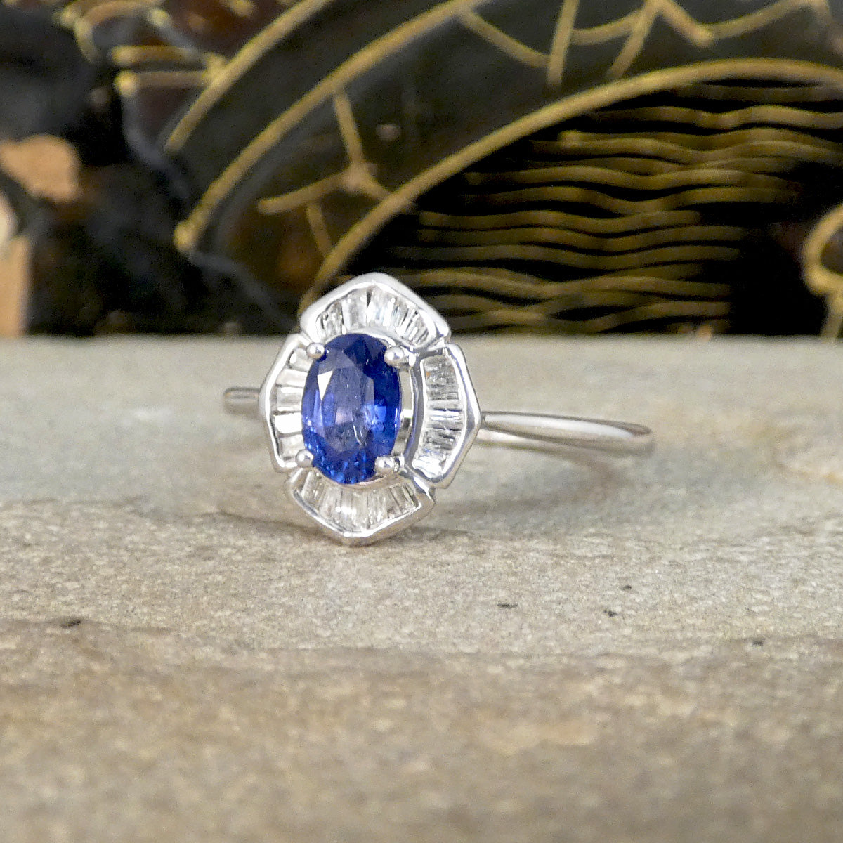 Front view of a white gold ring featuring a vibrant oval blue sapphire centre stone, encircled by a unique halo of baguette-cut clear gemstones, set in an art deco-inspired design, displayed on a stone surface with a decorative background.