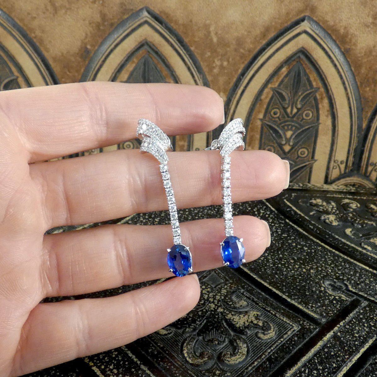 A hand holding the contemporary Sri Lankan Sapphire and Diamond drop earrings, highlighting the stunning craftsmanship with shimmering diamonds and vibrant blue sapphires against a decorative background.