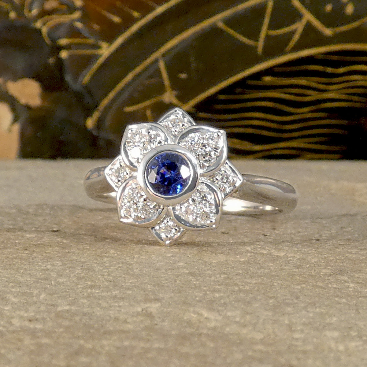 A flower cluster ring featuring a Ceylon Sapphire in the centre in a rub over bezel setting with a petal shaped Diamond clusters all made in Platinum.