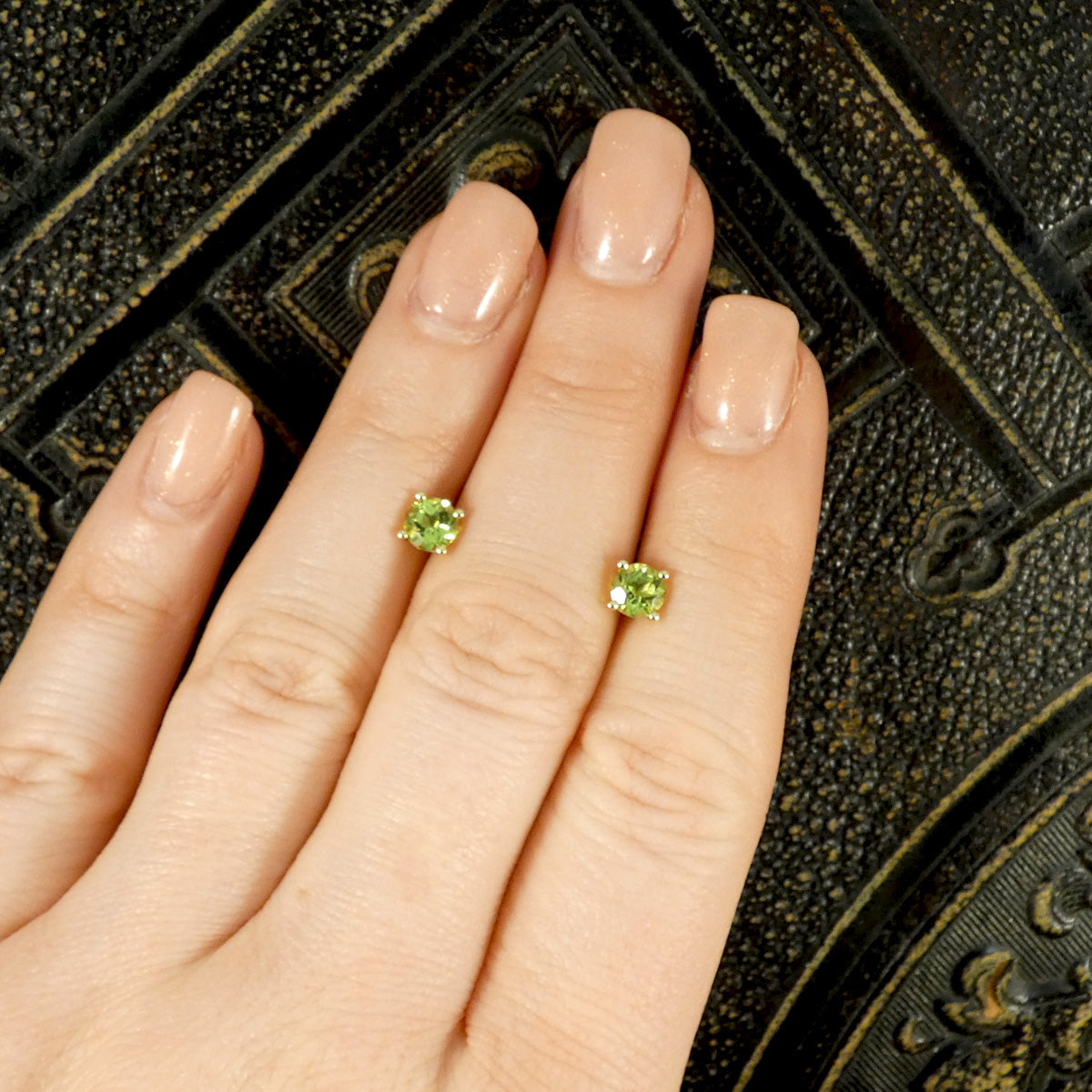 Four-claw peridot stud earrings crafted in 9ct yellow gold, featuring vibrant round-cut green gemstones with a classic and elegant design sat on hand for size ratio.