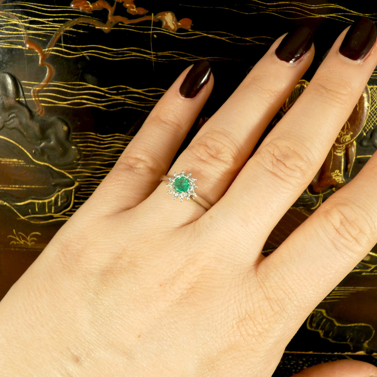 Emerald and diamond flower cluster ring in platinum displayed on a hand, highlighting its vibrant emerald centre piece and sparkling diamond halo.