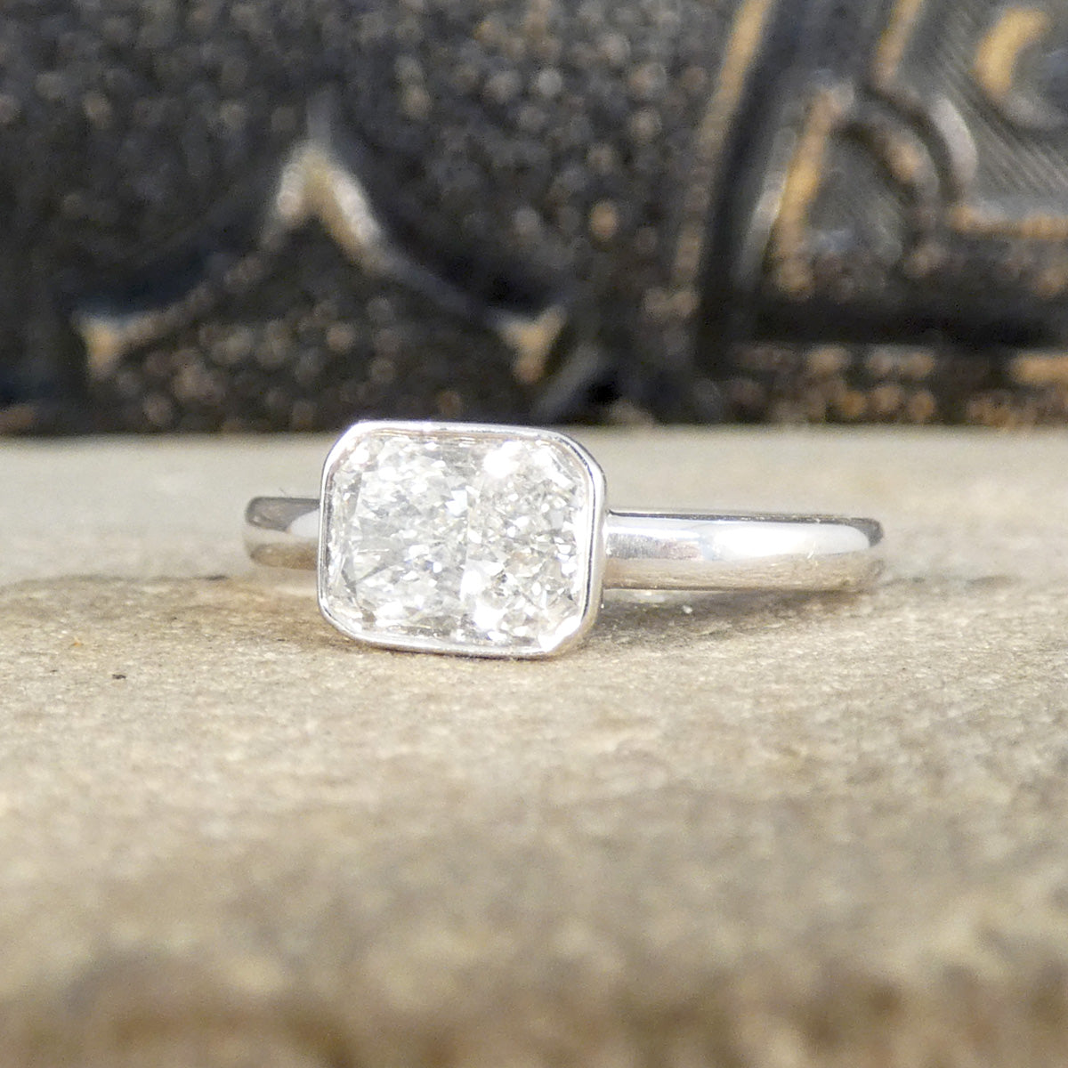 Radiant cut bezel set solitaire diamond ring in 18ct white gold, featuring a bright G-colour diamond independently assessed with a calculated weight of 1.29ct, displayed against a textured surface and ornate background.