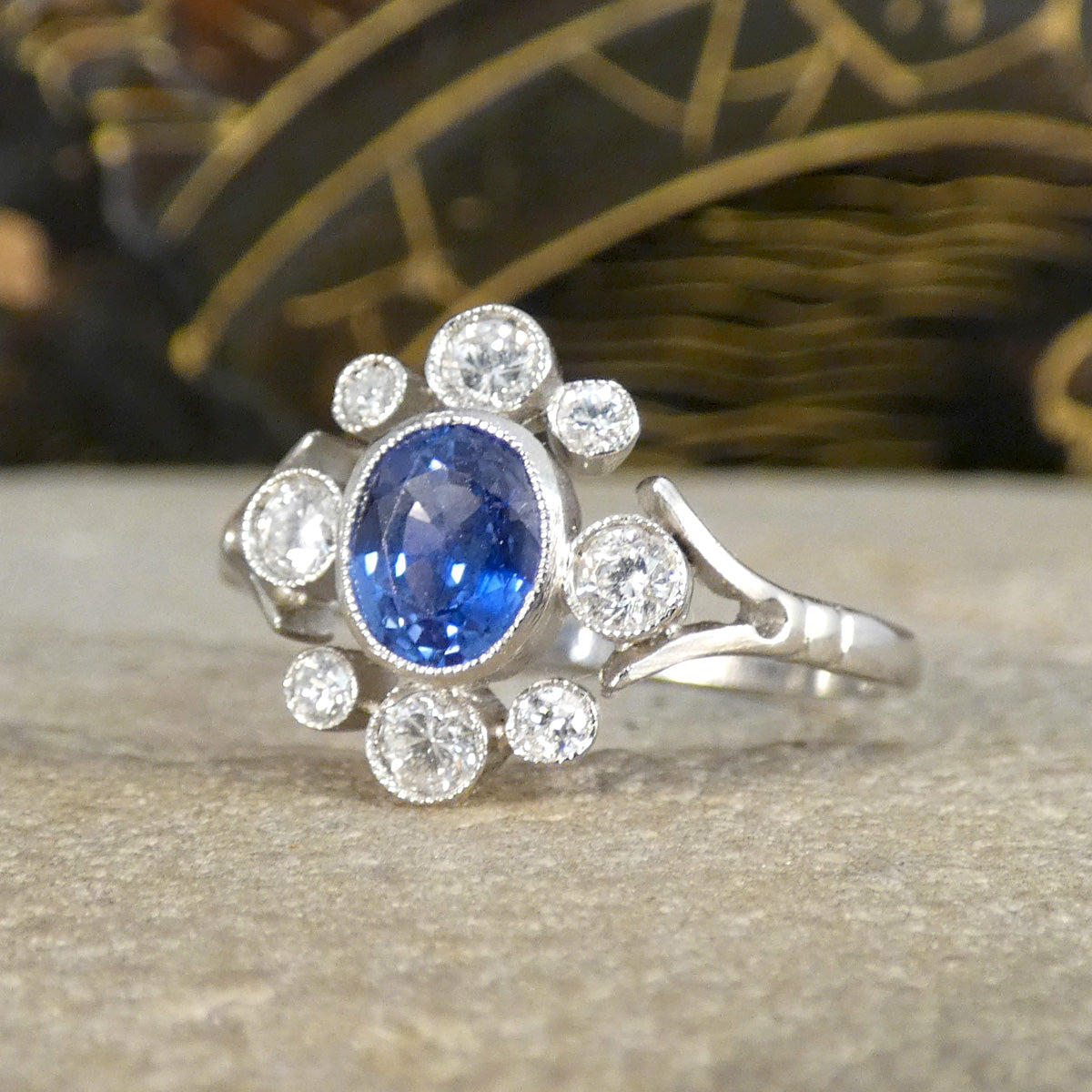 Angled view of a 0.80ct Sapphire and Diamond ring in platinum, showcasing the rich blue oval sapphire encircled by sparkling round diamonds in an elegant deco-style cluster, set on a delicate split shank.