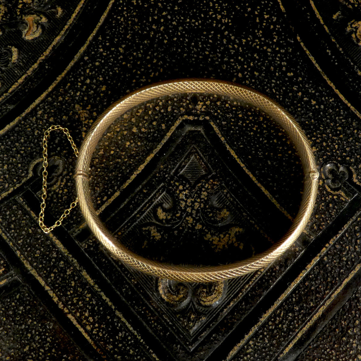 Vintage 9ct yellow gold hollowed bangle with a detailed textured pattern, featuring a safety chain, displayed on an ornate black and gold decorative surface. The perfect gift.