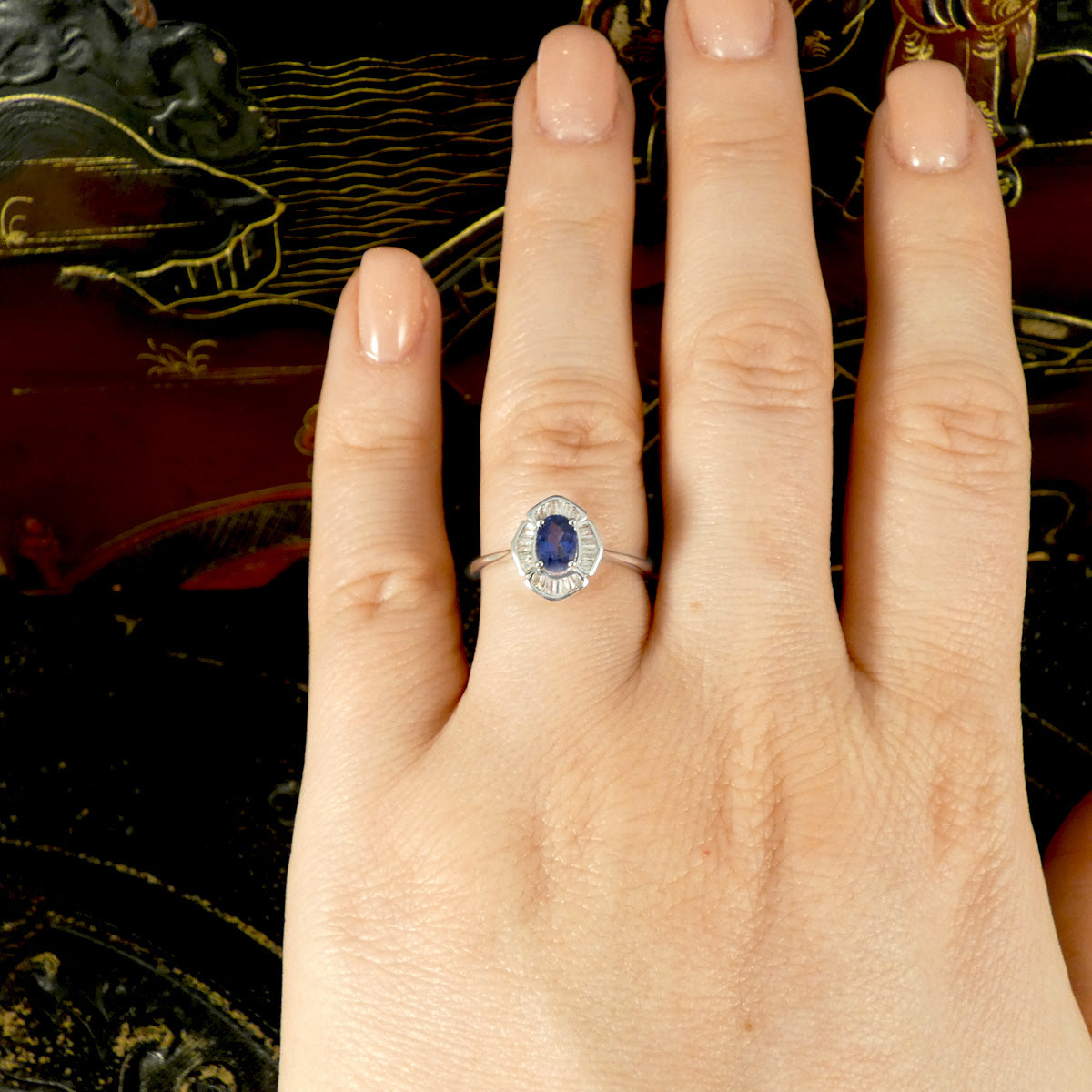 Hand wearing a white gold ring with an oval blue sapphire centre stone, framed by a halo of baguette-cut clear gemstones in an art deco-inspired design, displayed against a decorative background, highlighting the ring’s elegance and scale on the finger.