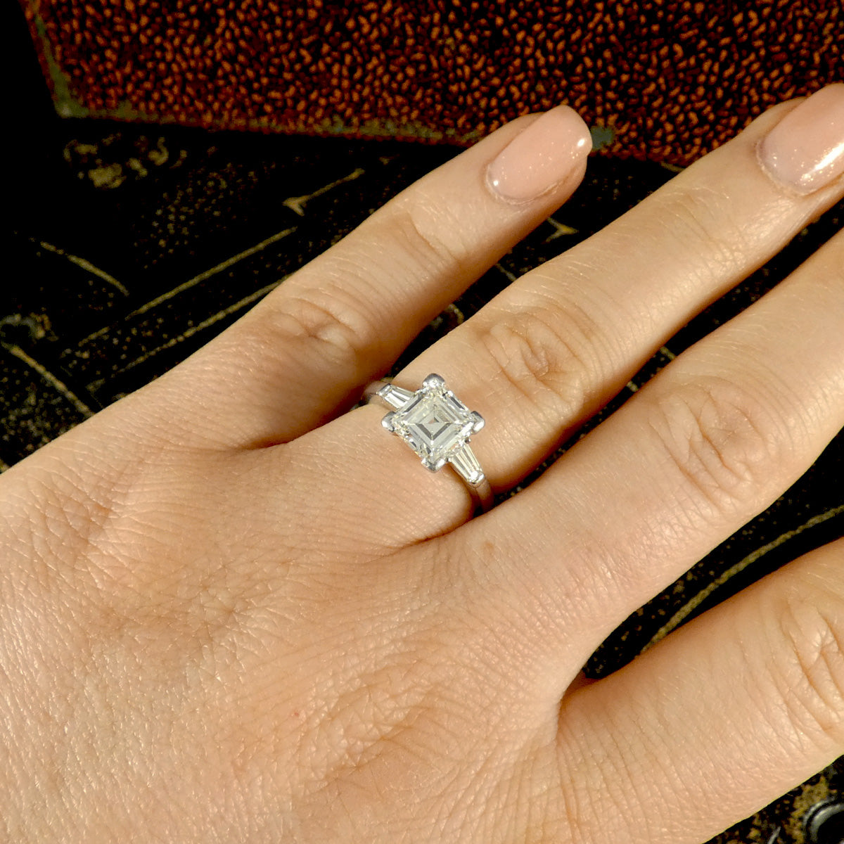 Hand wearing an elegant 18-carat white gold ring with a total diamond weight of 2.00 carats, featuring a 1.75ct square emerald-cut centre diamond accented by two tapered baguette diamonds, highlighting its refined and timeless three-stone design. Worn on engagement ring finger.