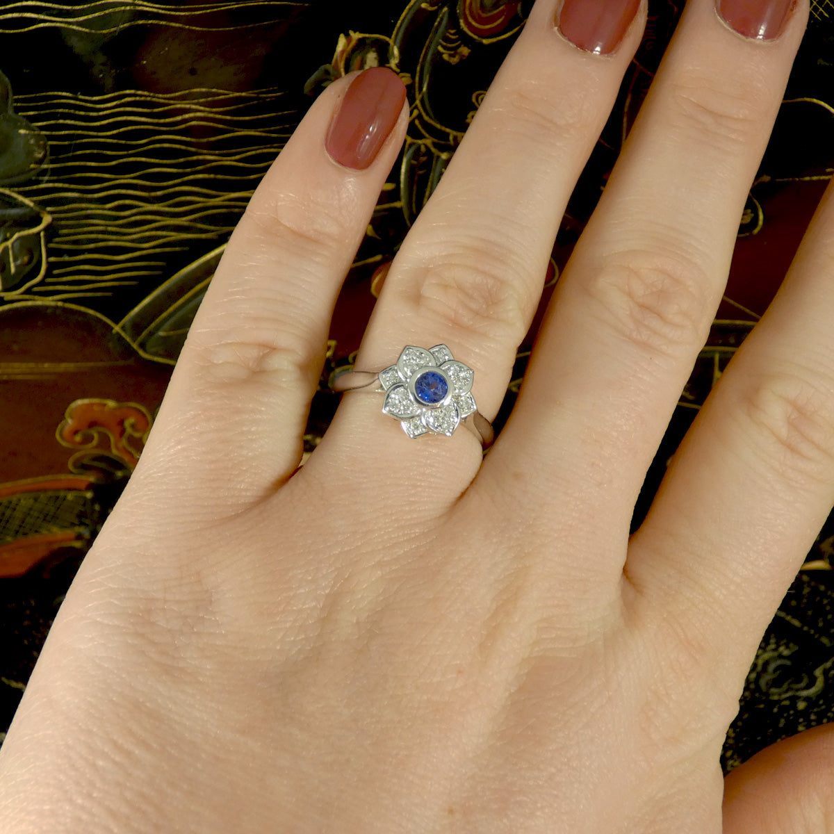 A flower cluster ring featuring a Ceylon Sapphire in the centre in a rub over bezel setting with a petal shaped Diamond clusters all made in Platinum. Shown on hand to see how the ring would look on the ring finger.