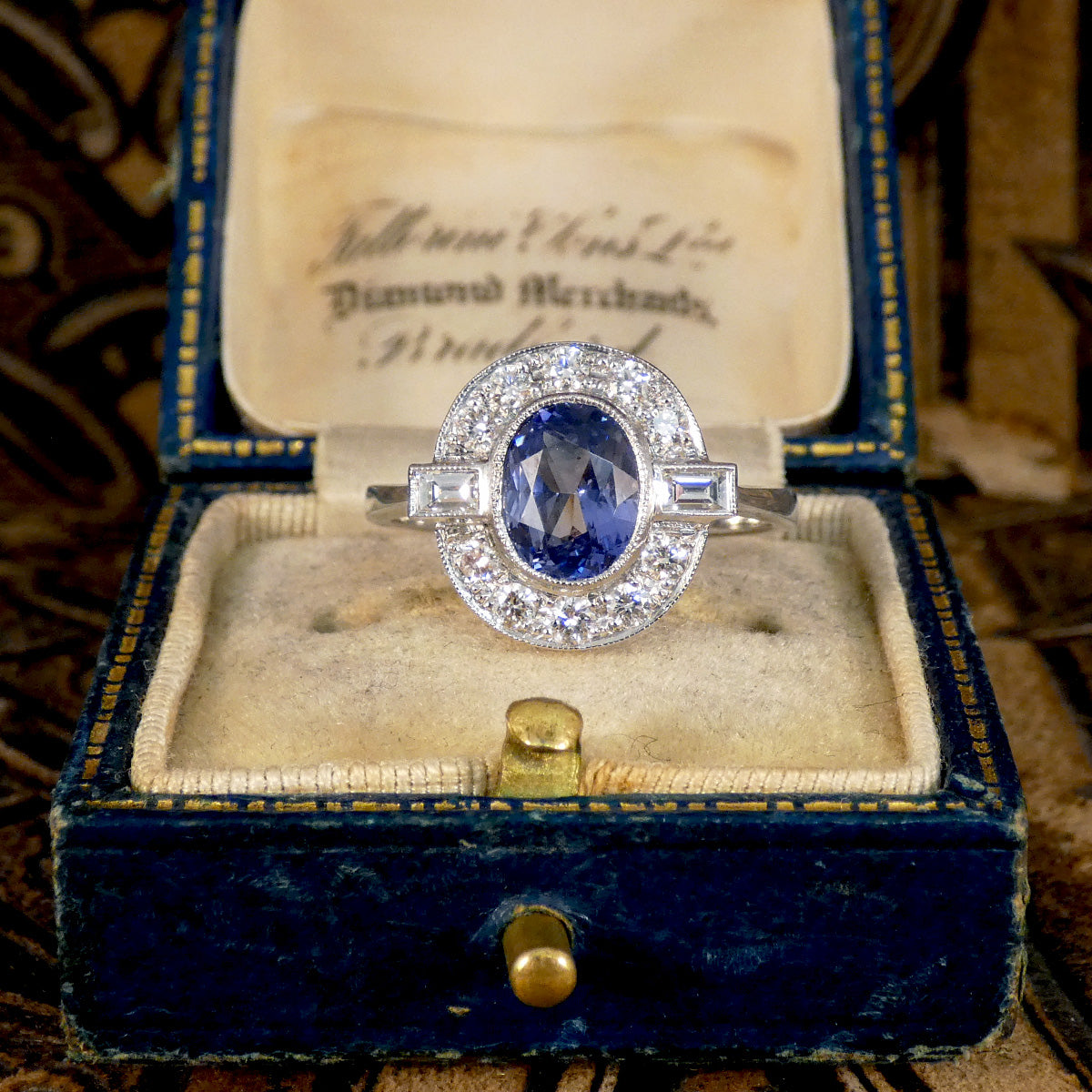 An oval cut Sapphire with a rub over bezel setting surrounded by Brilliant and Baguette cut Diamonds clustering the velvet blue centre stone. A true are deco style ring, a beautiful vintage classic. Shown in vintage box.