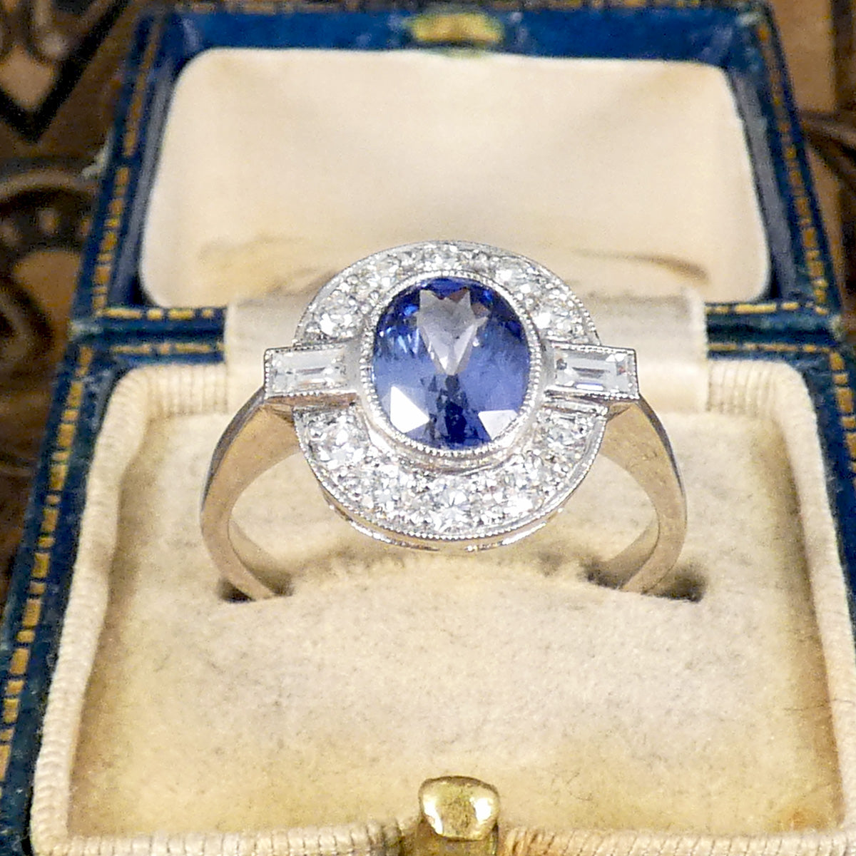 An oval cut Sapphire with a rub over bezel setting surrounded by Brilliant and Baguette cut Diamonds clustering the velvet blue centre stone. A true are deco style ring, a beautiful vintage classic.