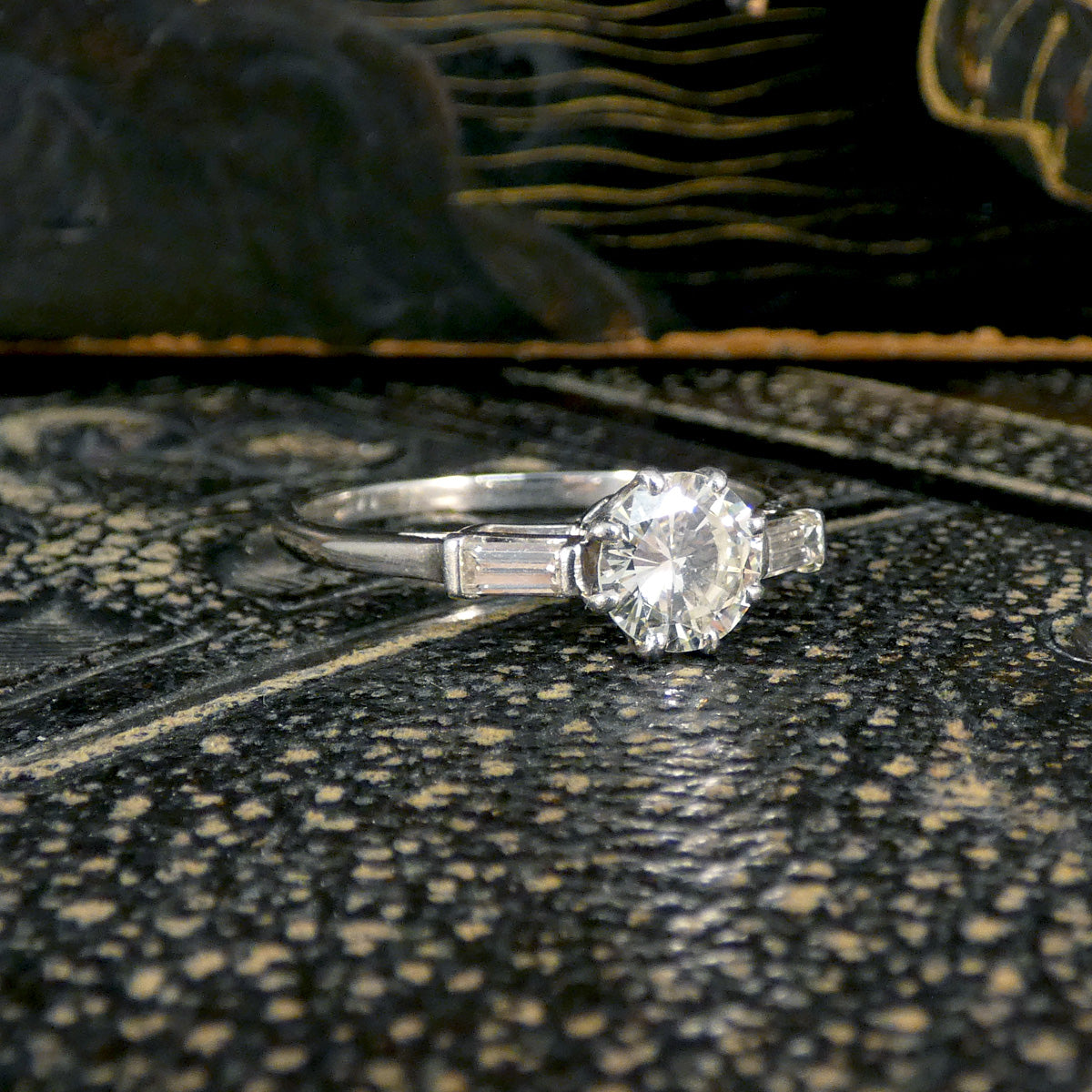 Platinum engagement ring featuring a 0.90-carat early brilliant-cut centre diamond, complemented by 0.10-carat baguette-cut diamonds on each side. The ring's sleek design and radiant sparkle are showcased on a textured surface, highlighting its timeless elegance.