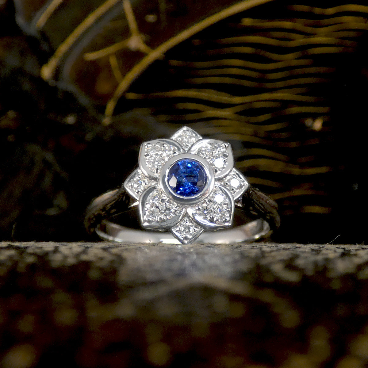 A flower cluster ring featuring a Ceylon Sapphire in the centre in a rub over bezel setting with a petal shaped Diamond clusters all made in Platinum. Really bright blue and perfect gift.