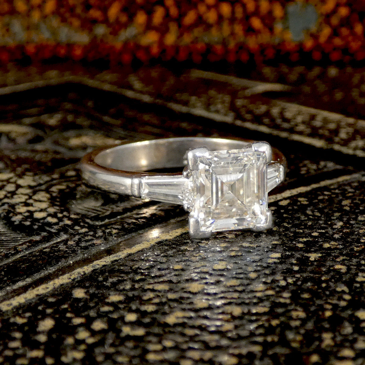 Classic three-stone ring featuring a square emerald-cut centre diamond flanked by tapered baguette side stones, set in 18-carat white gold with a total diamond weight of 2.00 carats, displayed on a textured, vintage-inspired surface for a timeless and sophisticated appeal.