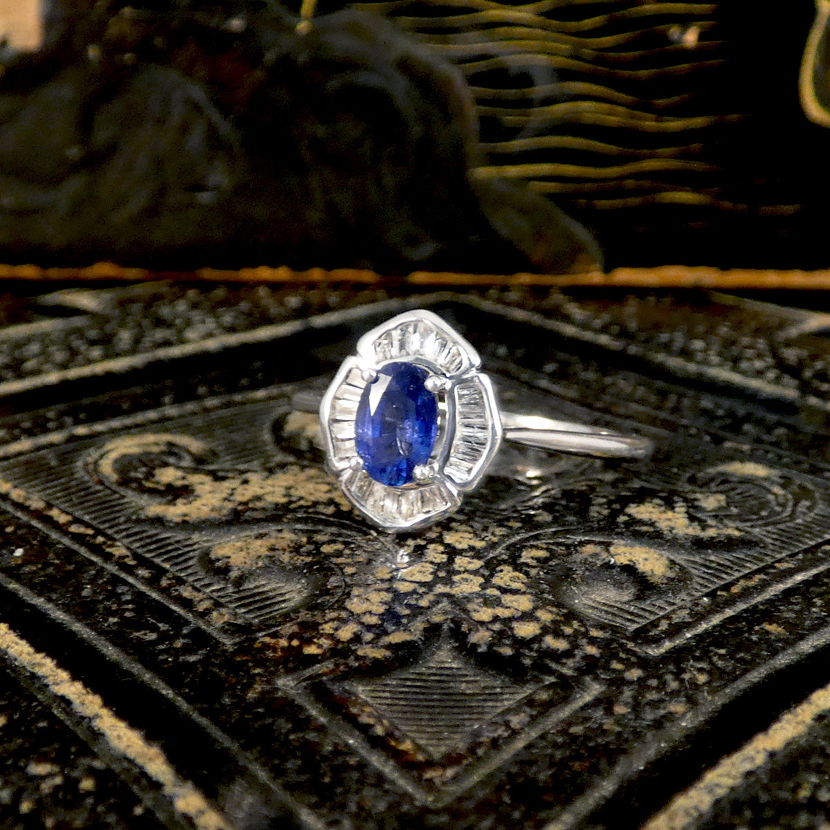 18ct White Gold ring featuring an oval 0.66ct blue sapphire centre stone, surrounded by a halo of 0.13ct baguette-cut clear gemstones in a unique, vintage-inspired setting, displayed on an ornate, textured surface that enhances the ring’s elegance.