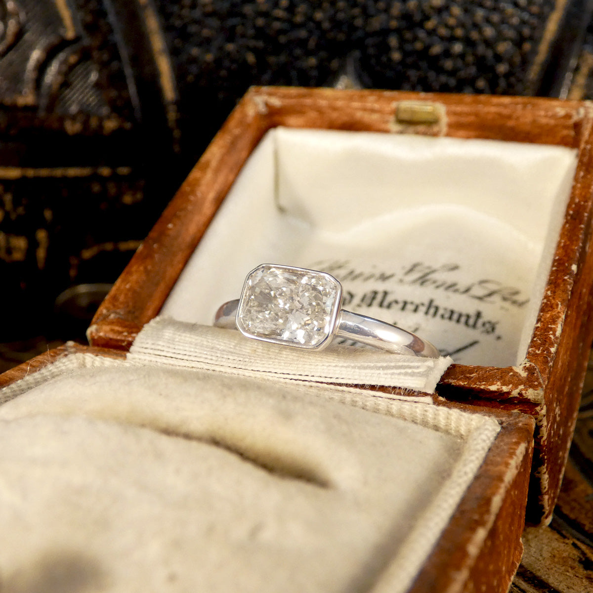 Radiant cut east-west bezel set solitaire engagement ring in 18ct white gold, featuring a G-colour diamond with a calculated weight of 1.29ct, elegantly placed in a vintage wooden ring box with a cream fabric interior.