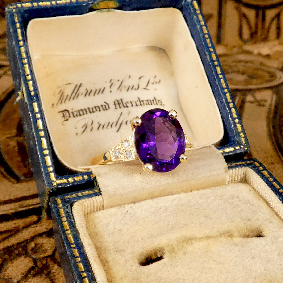 Bright and vibrant amethyst ring in a four claw setting with diamond set shoulders in 9ct yellow gold. shown in vintage box mirroring the aesthetic of the ring.