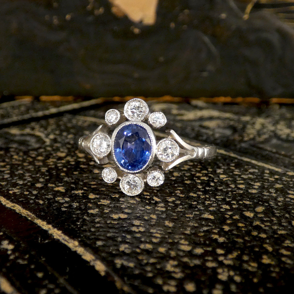 Platinum Sapphire and Diamond ring, elegantly showcased against a textured dark surface, featuring a striking blue sapphire surrounded by brilliant round diamonds in a timeless Art Deco-inspired design.