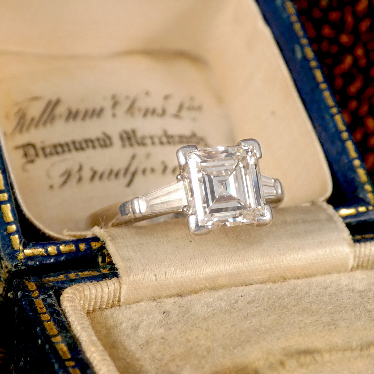 Vintage-inspired three-stone diamond ring featuring a square emerald-cut centre stone with tapered baguette side stones, set in 18-carat white gold with a total diamond weight of 2.00 carats, elegantly displayed in a classic velvet-lined jewellery box, enhancing its timeless sophistication.