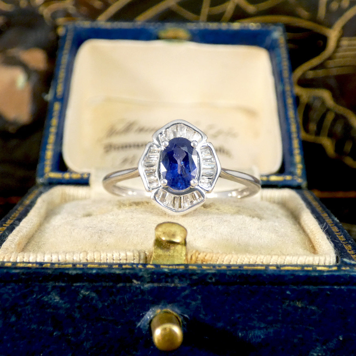 White Gold ring with an oval blue sapphire surrounded by baguette-cut clear gemstones, displayed in an elegant jewellery box with a velvet interior, adding a vintage touch to the sophisticated presentation.