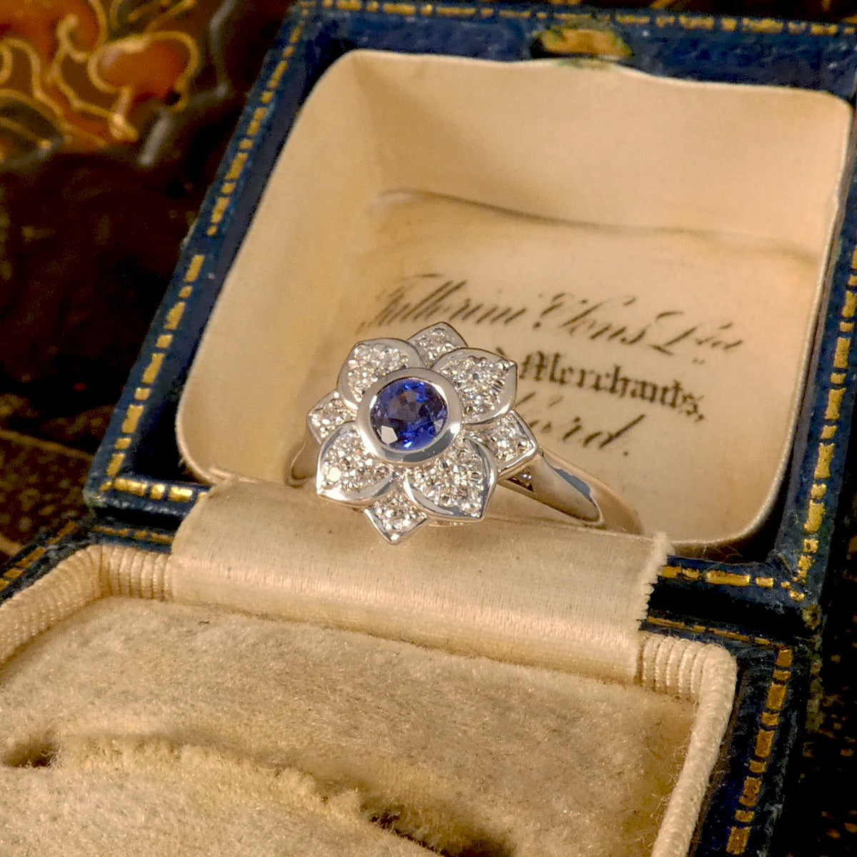 A flower cluster ring featuring a Ceylon Sapphire in the centre in a rub over bezel setting with a petal shaped Diamond clusters all made in Platinum. Displayed in vintage box.