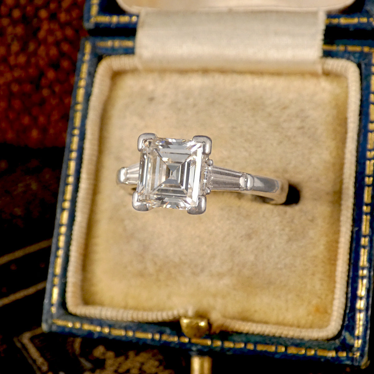 Exquisite three-stone diamond ring in 18-carat white gold, featuring a square emerald-cut center diamond with exceptional clarity and brilliance, flanked by two tapered baguette diamonds. Displayed in a vintage velvet-lined box, this ring captures timeless beauty and elegance.