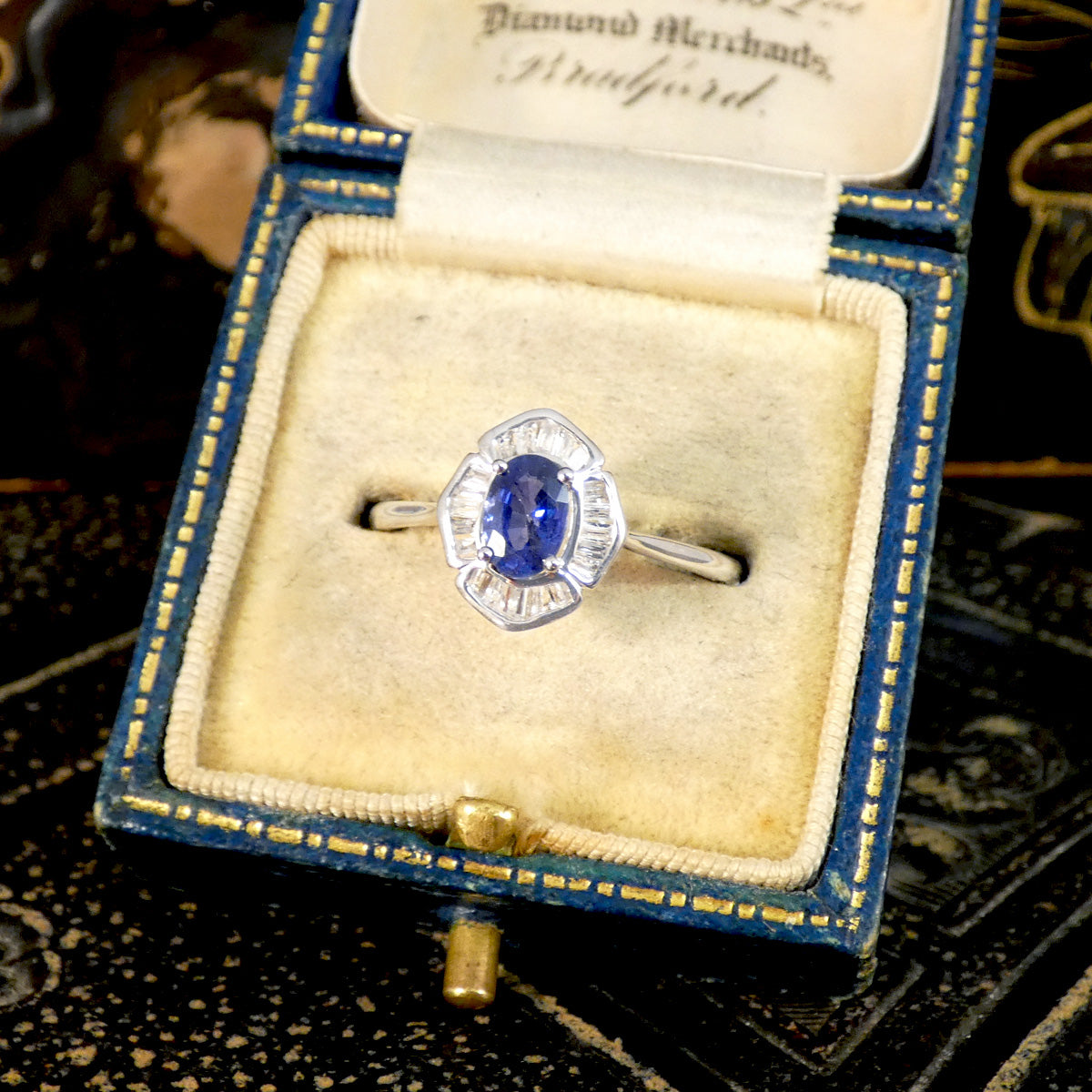 White Gold ring with a 0.66ct oval blue sapphire centre piece, framed by baguette-cut clear gemstones in mix between an art deco and modern style setting, elegantly placed in a vintage-inspired blue velvet jewellery box with gold accents, enhancing its timeless appeal.