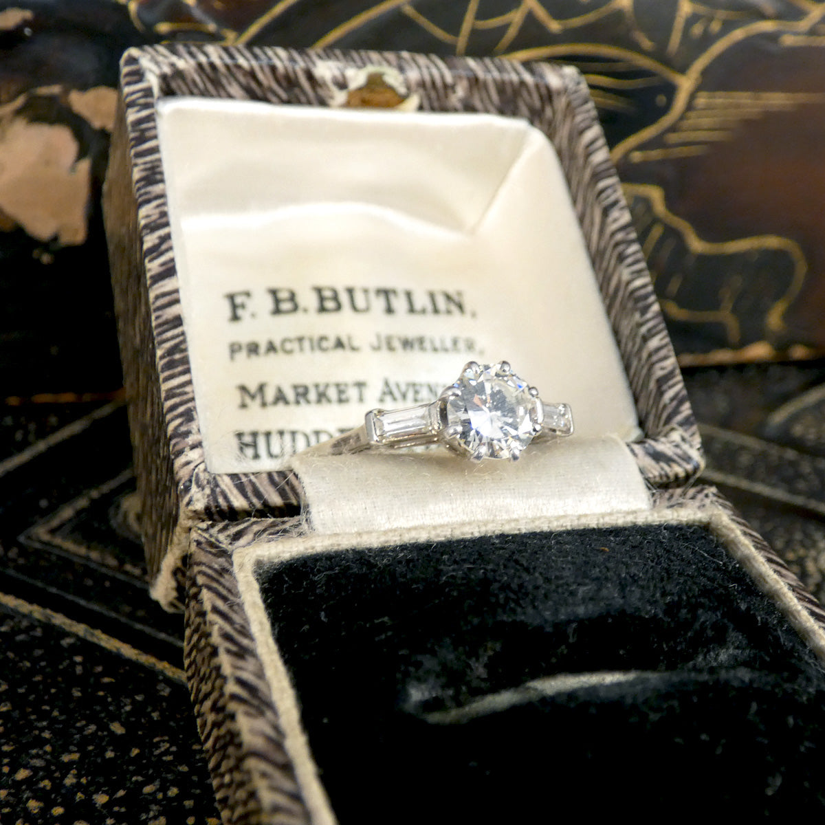 Platinum engagement ring featuring a 0.90-carat early brilliant-cut centre diamond and 0.10-carat baguette-cut diamonds on each side, presented in a vintage jewellery box from F.B. Butlin. This timeless and elegant design offers exceptional brilliance, perfect for a classic proposal.